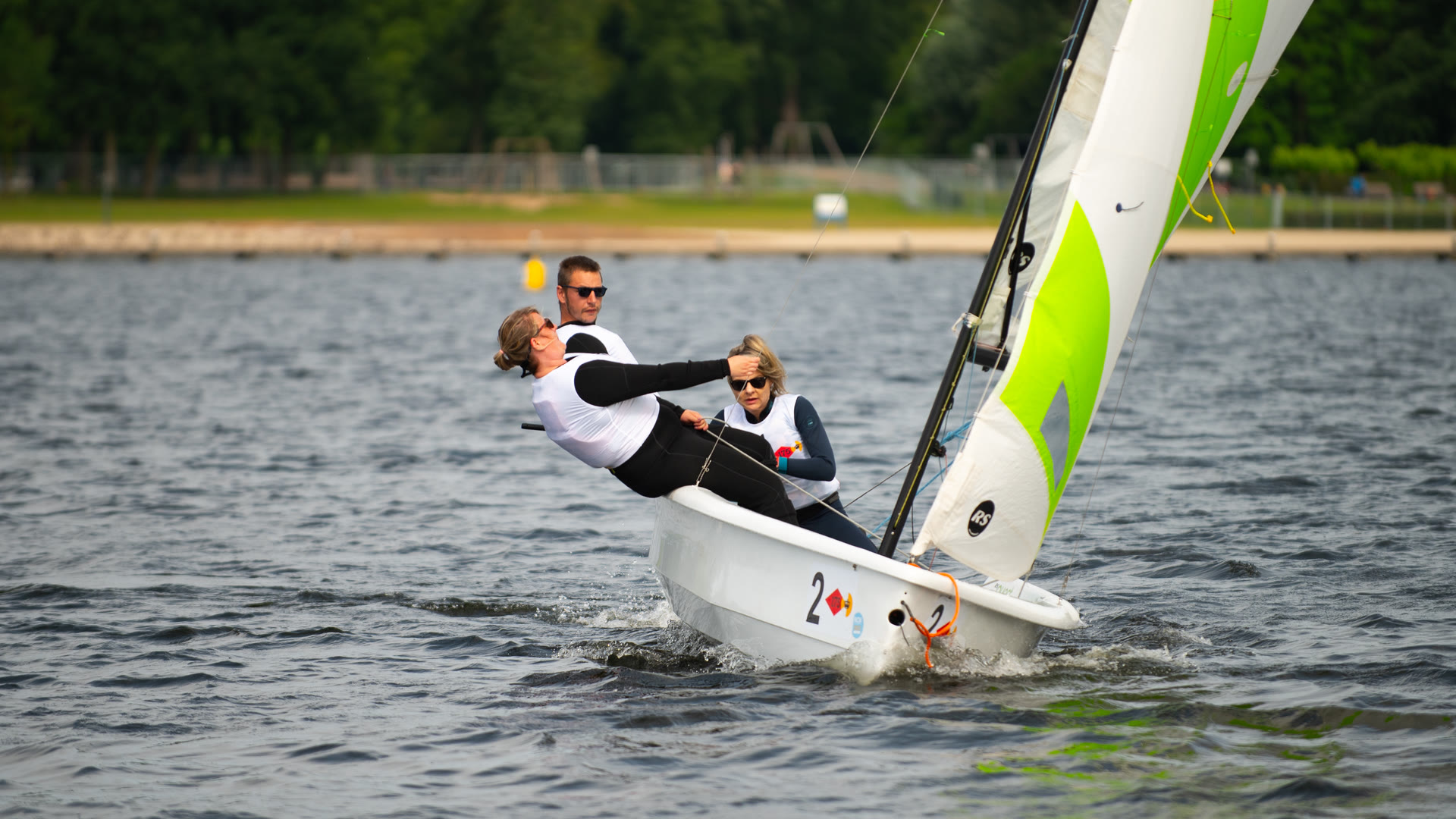 Een driekoppige bemanning kruist aan de wind in de volle zon, een van de zeilers hangt ver buitenboord om de boot vlak te houden.